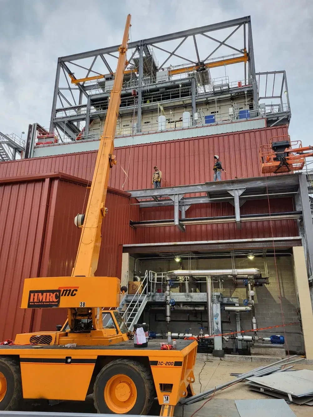 An orange crane lifts a structure near a large industrial building, with workers on the roof.