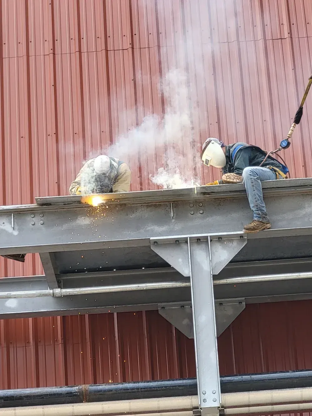 Two welders working on a metal beam, sparks and smoke visible; red building background.