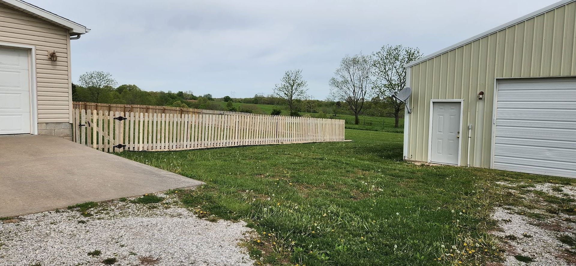 Gravel driveway beside beige garages and a wooden fence, with a grassy yard and overcast sky.