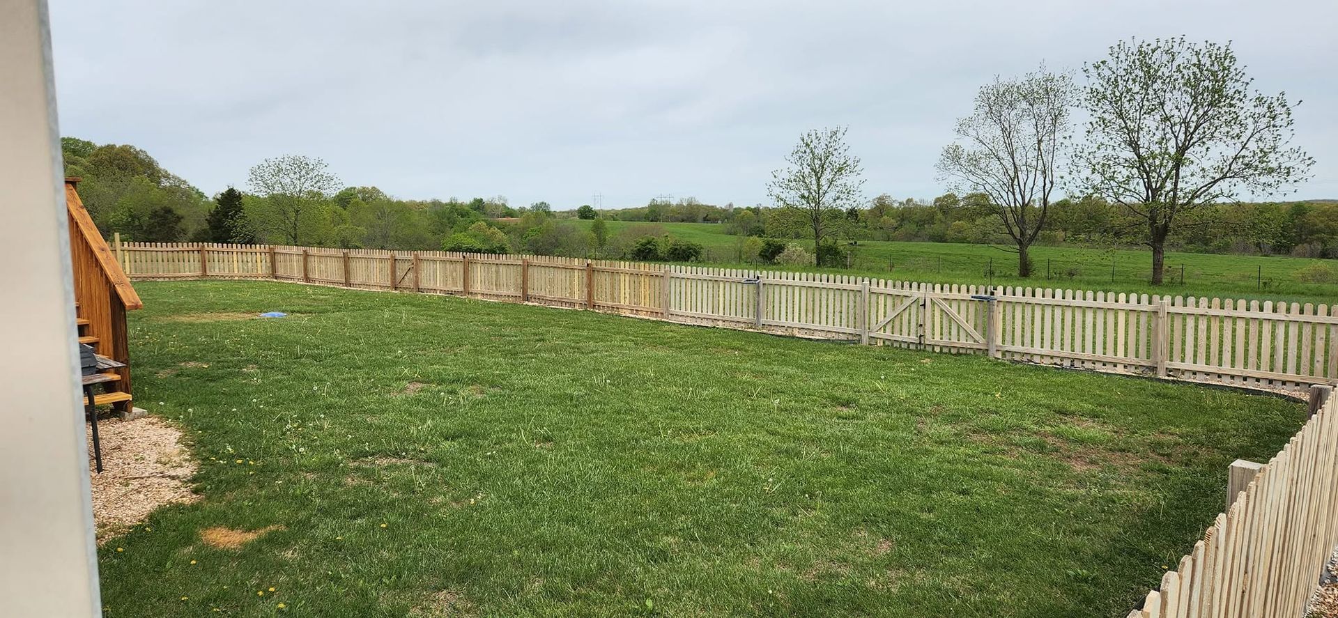 Large grassy backyard with a white fence, trees, and cloudy sky