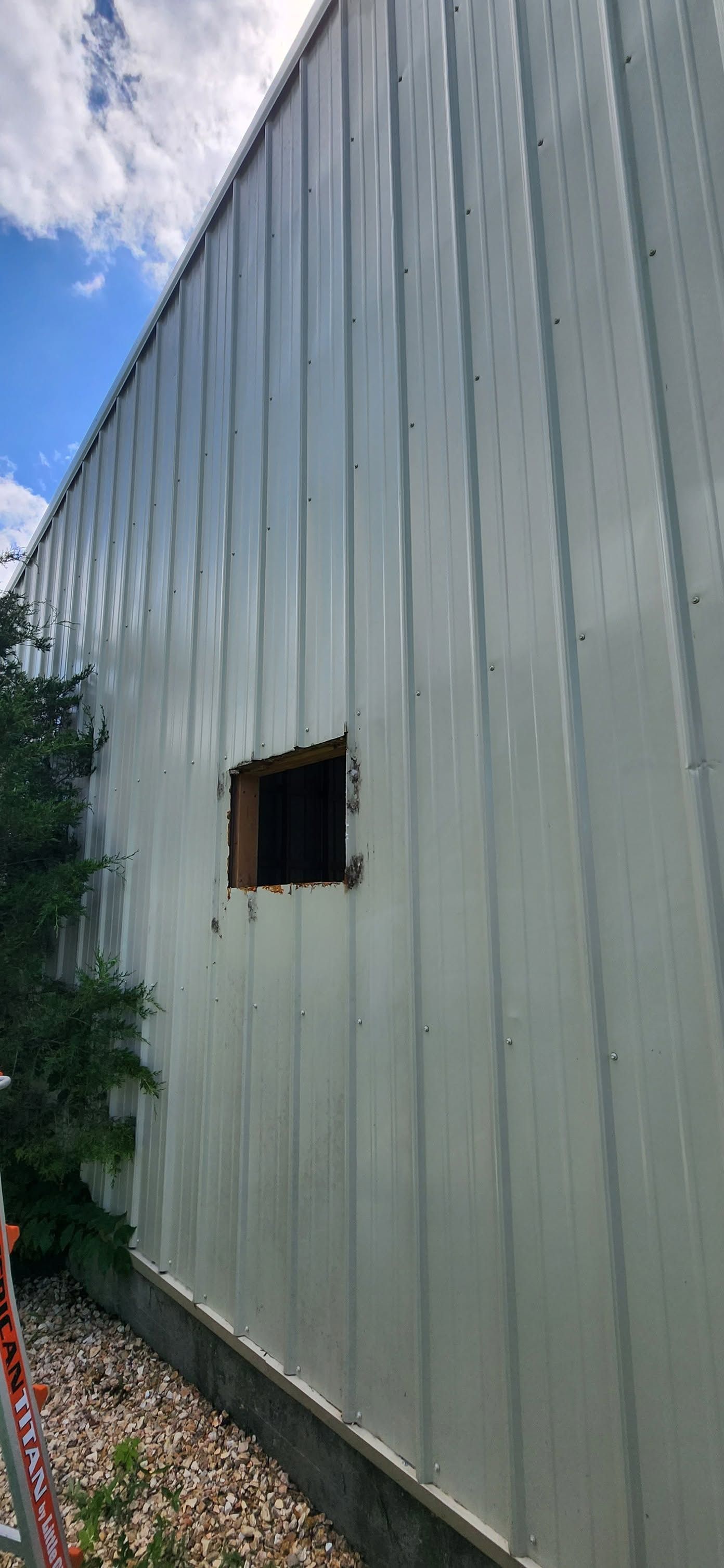 Tall light-gray corrugated metal building wall with a small square window and gravel below