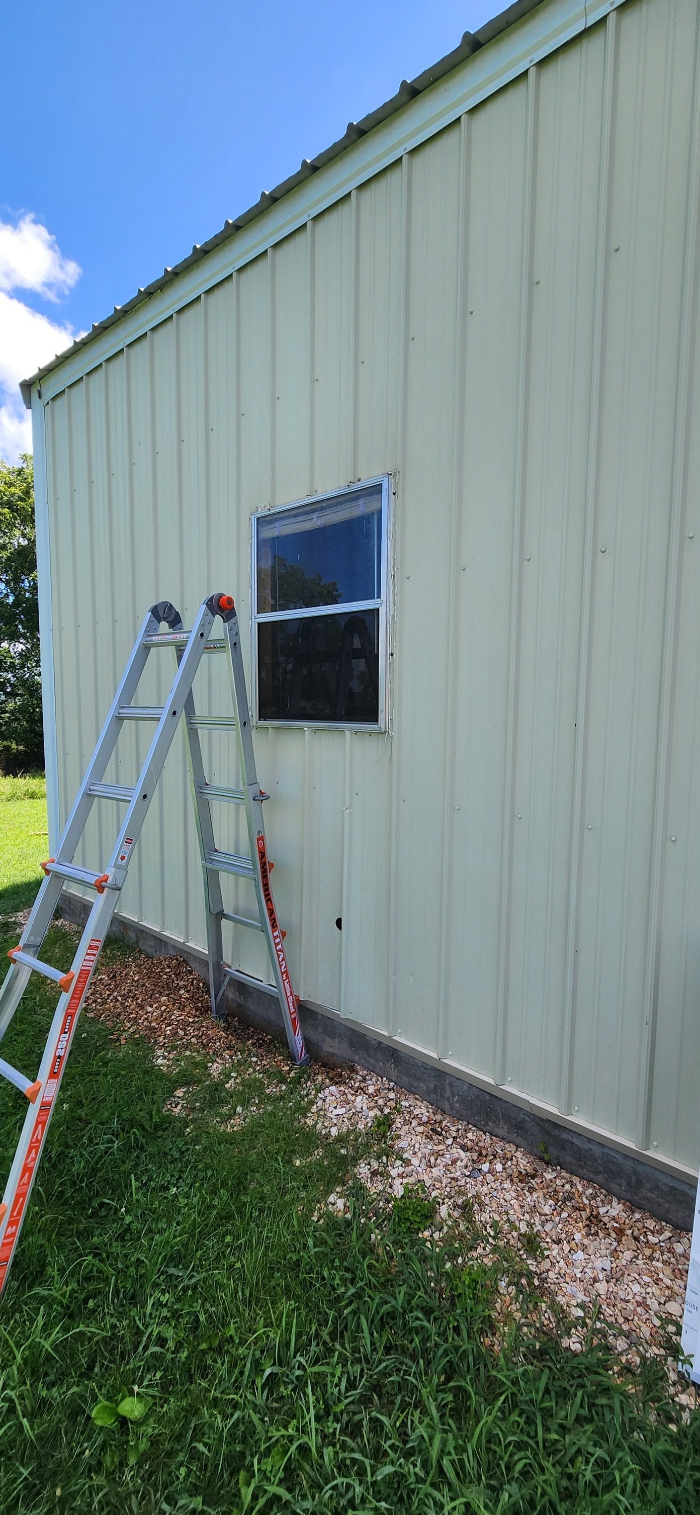 Ladder leaning against a pale green metal building beside a window with grass in front