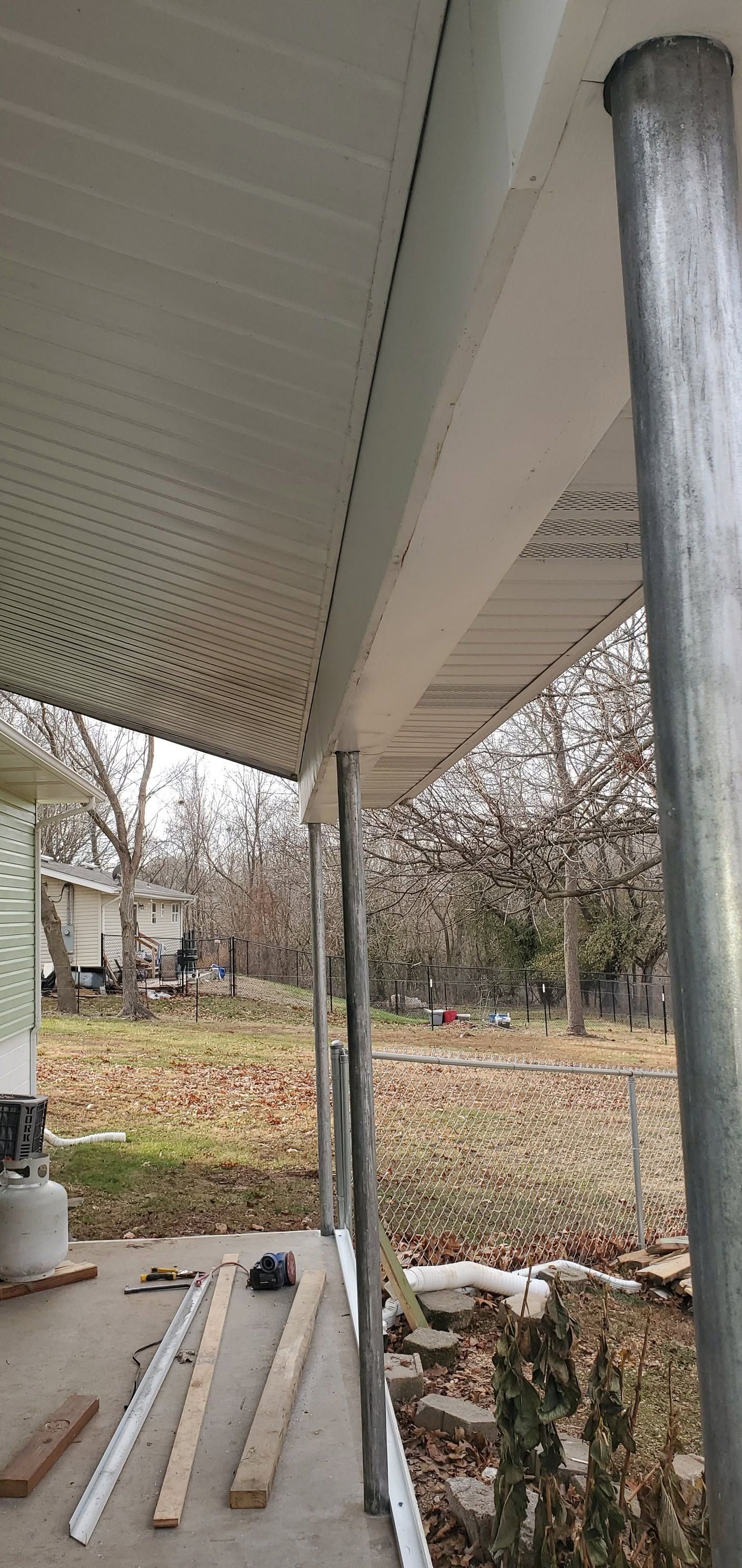 Under a porch roof, construction materials lie on the ground beside a white support column and a metal downspout.