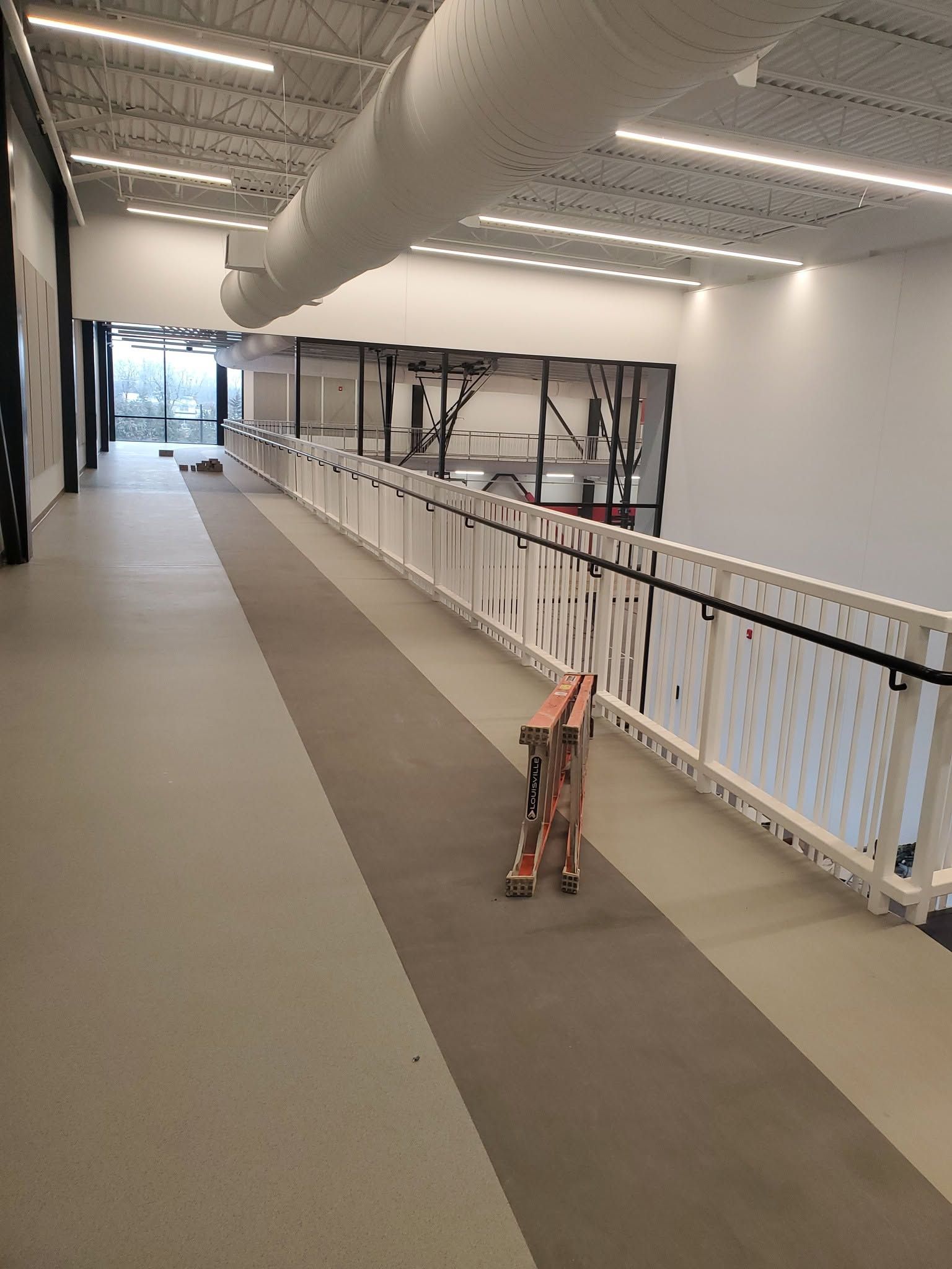 Long indoor hallway with a white railing, gray floor, and construction barriers along one side.