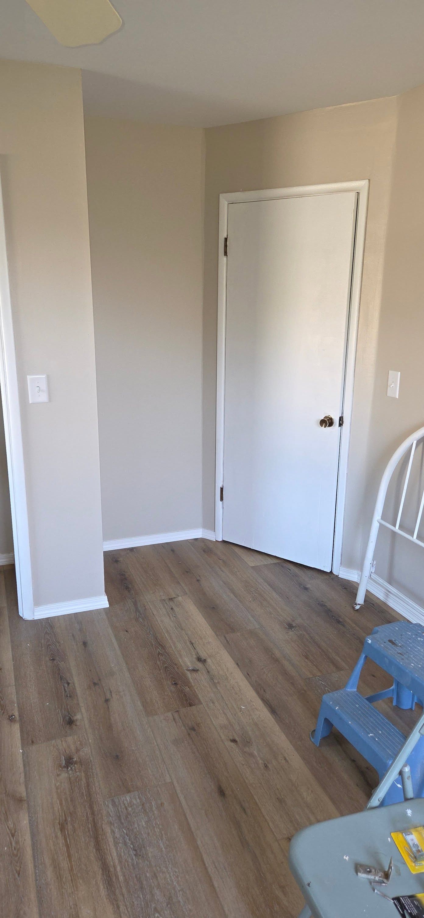 Empty beige room with white door, wood floor, and a blue ladder in the corner