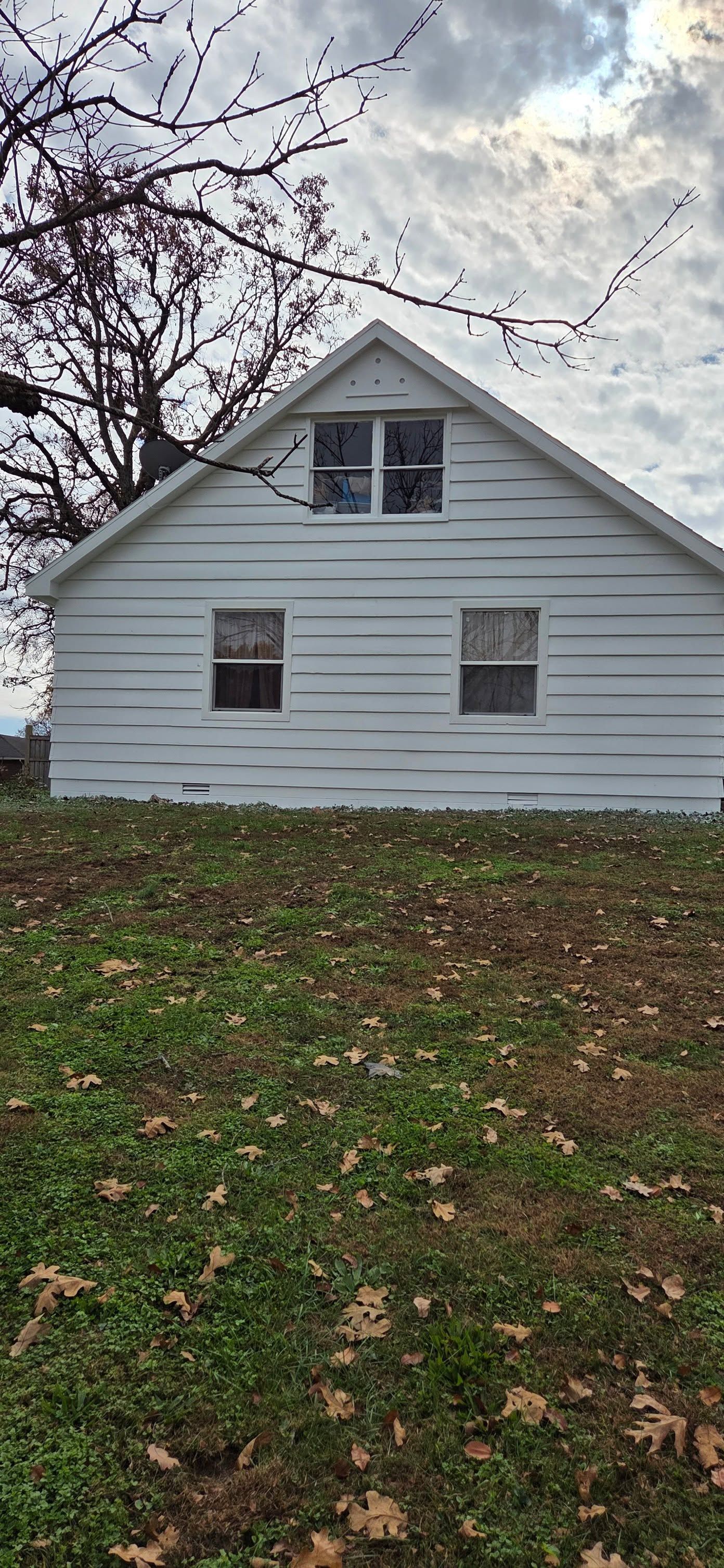 White house with a steep roof and dormer windows, seen from a leaf-covered yard under cloudy sky.