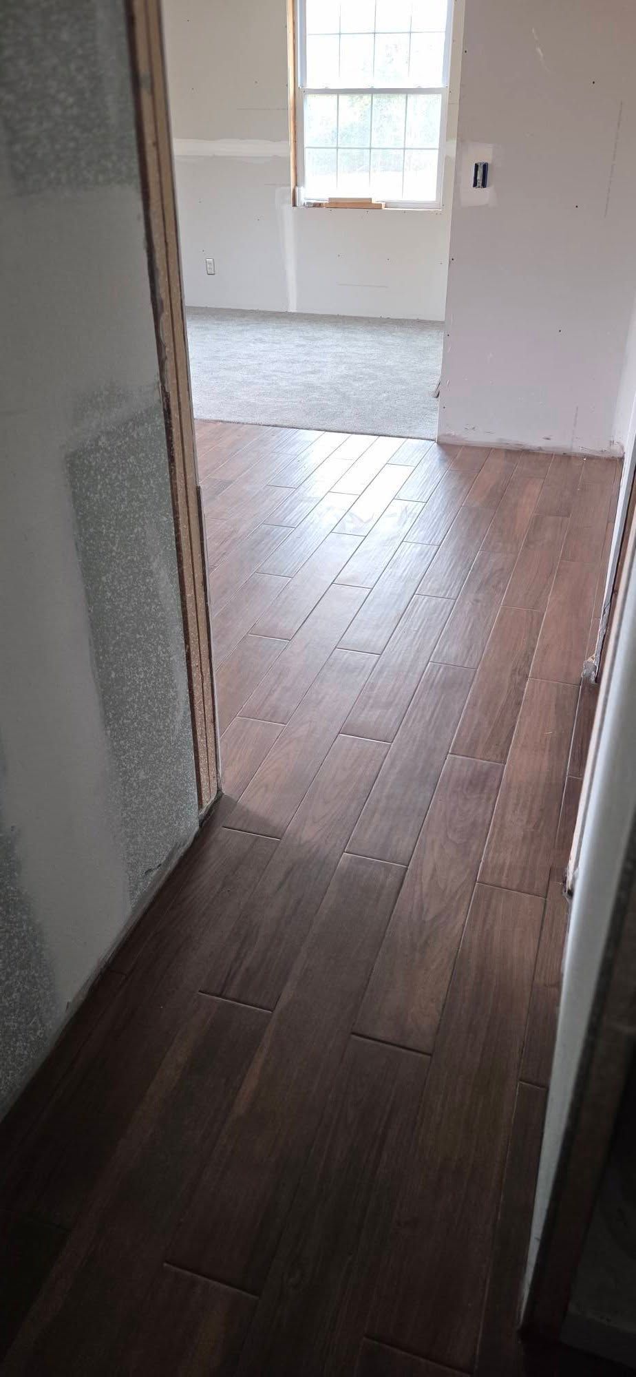 Narrow hallway with dark wood flooring leading to a bright room with white walls and a window.