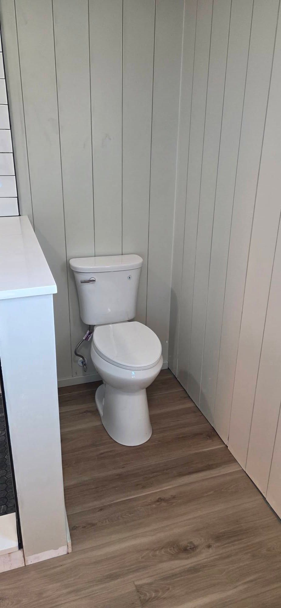 Small bathroom with a white toilet against light paneled walls and wood-look flooring.