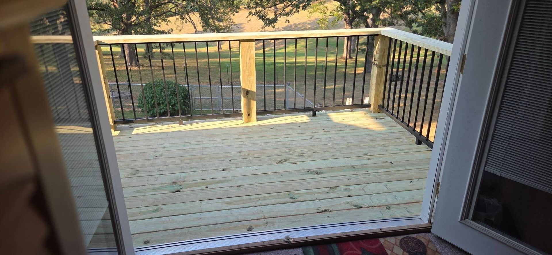 Wooden deck with black metal railing, viewed through an open doorway, overlooking trees and a grassy yard