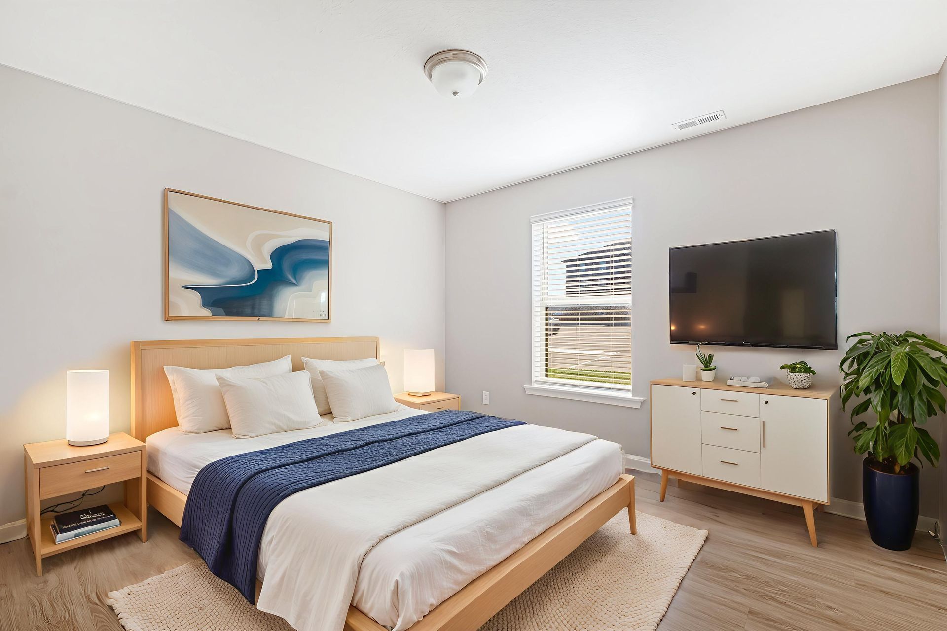 Bright bedroom with a neatly made bed, nightstand lamps, wall art, TV, and a window with blinds.