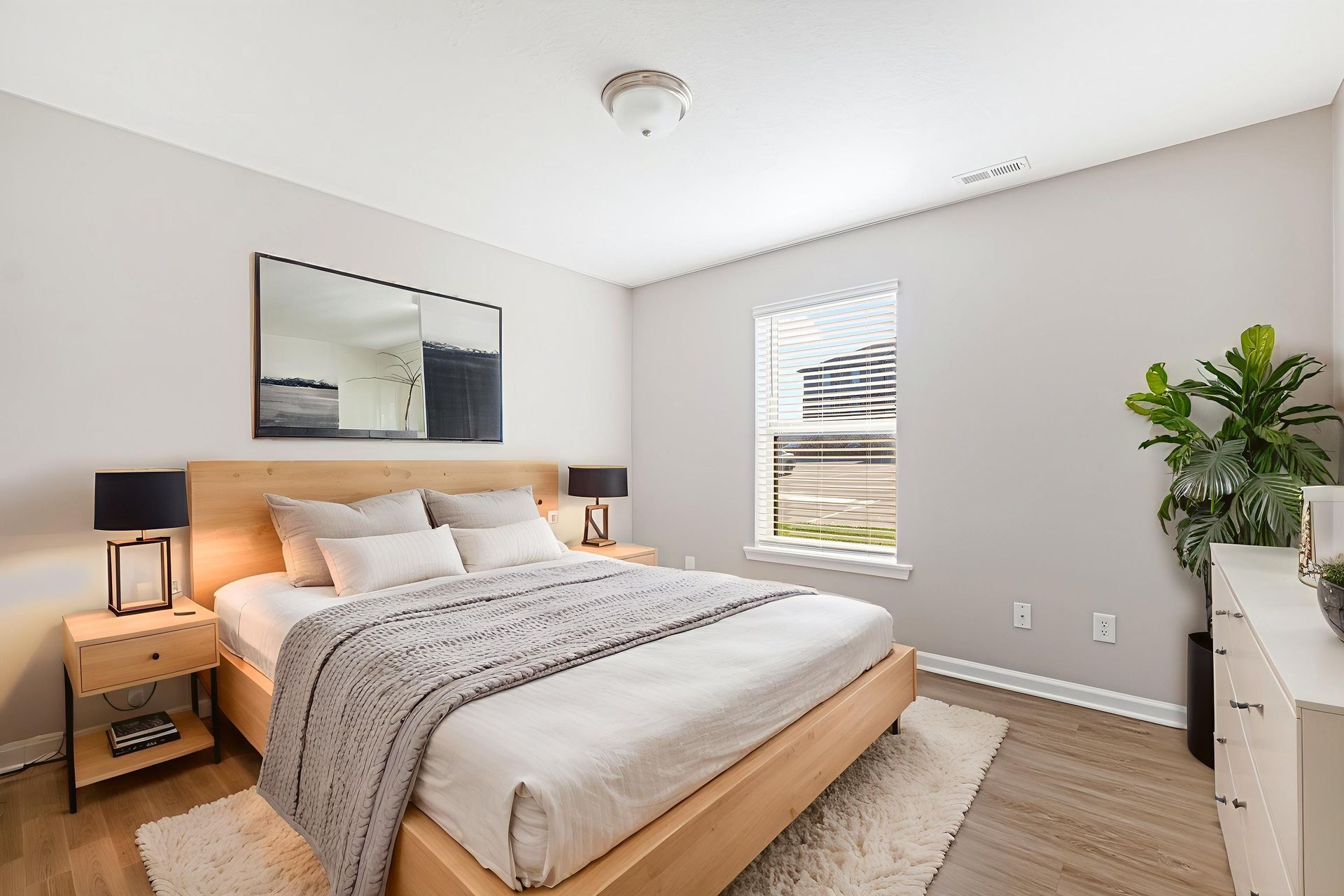 Bright bedroom with a made bed, nightstand lamps, mirror, window, and potted plant