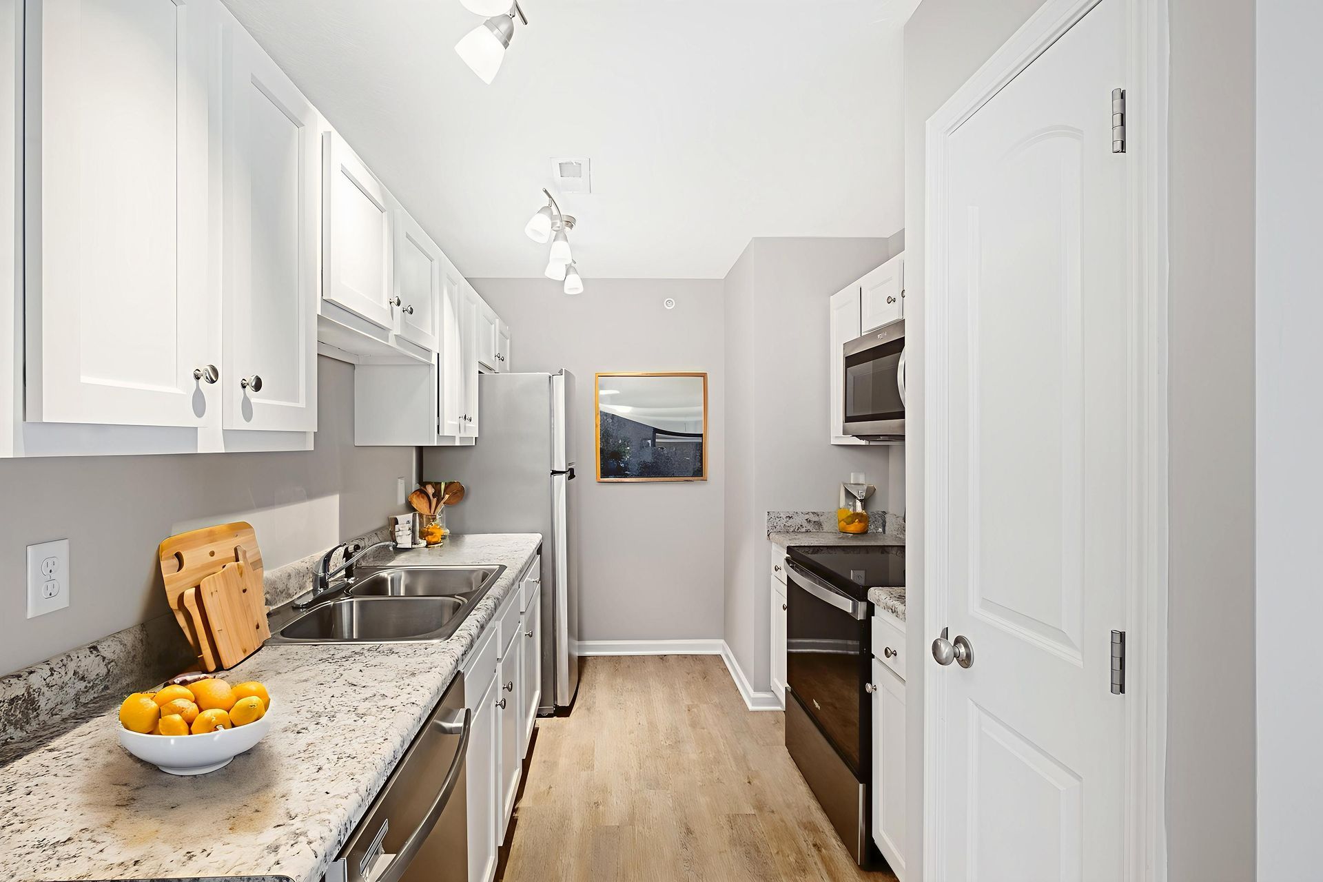 Bright narrow kitchen with white cabinets, granite counters, stainless appliances, and wood flooring.