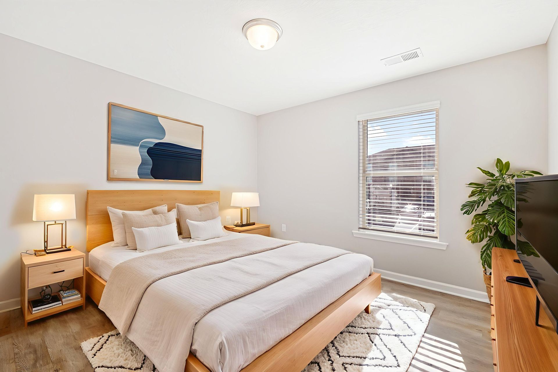 Bright bedroom with a neatly made bed, bedside lamps, wall art, and a sunlit window.