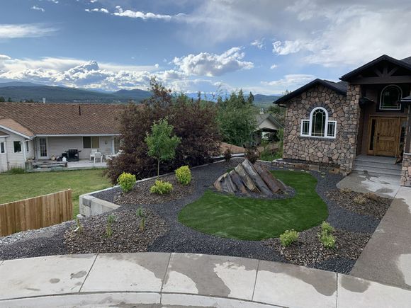 Suburban front yard with rock garden, green lawn, stone house, and mountain view under cloudy sky