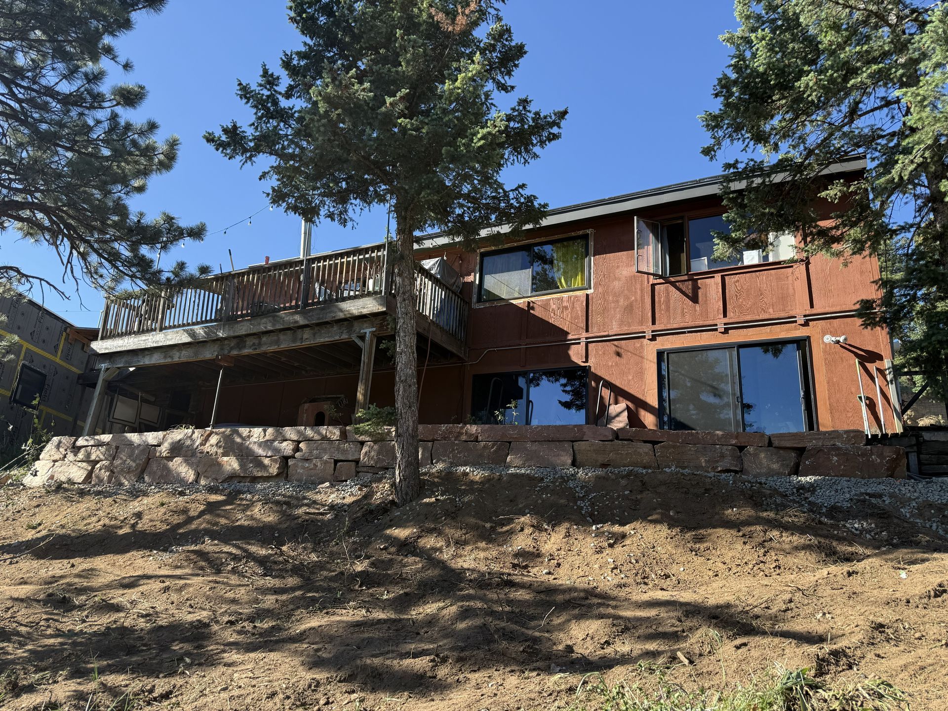 Two-story brick house on a hillside with tall pine trees and a wooden deck under a clear sky
