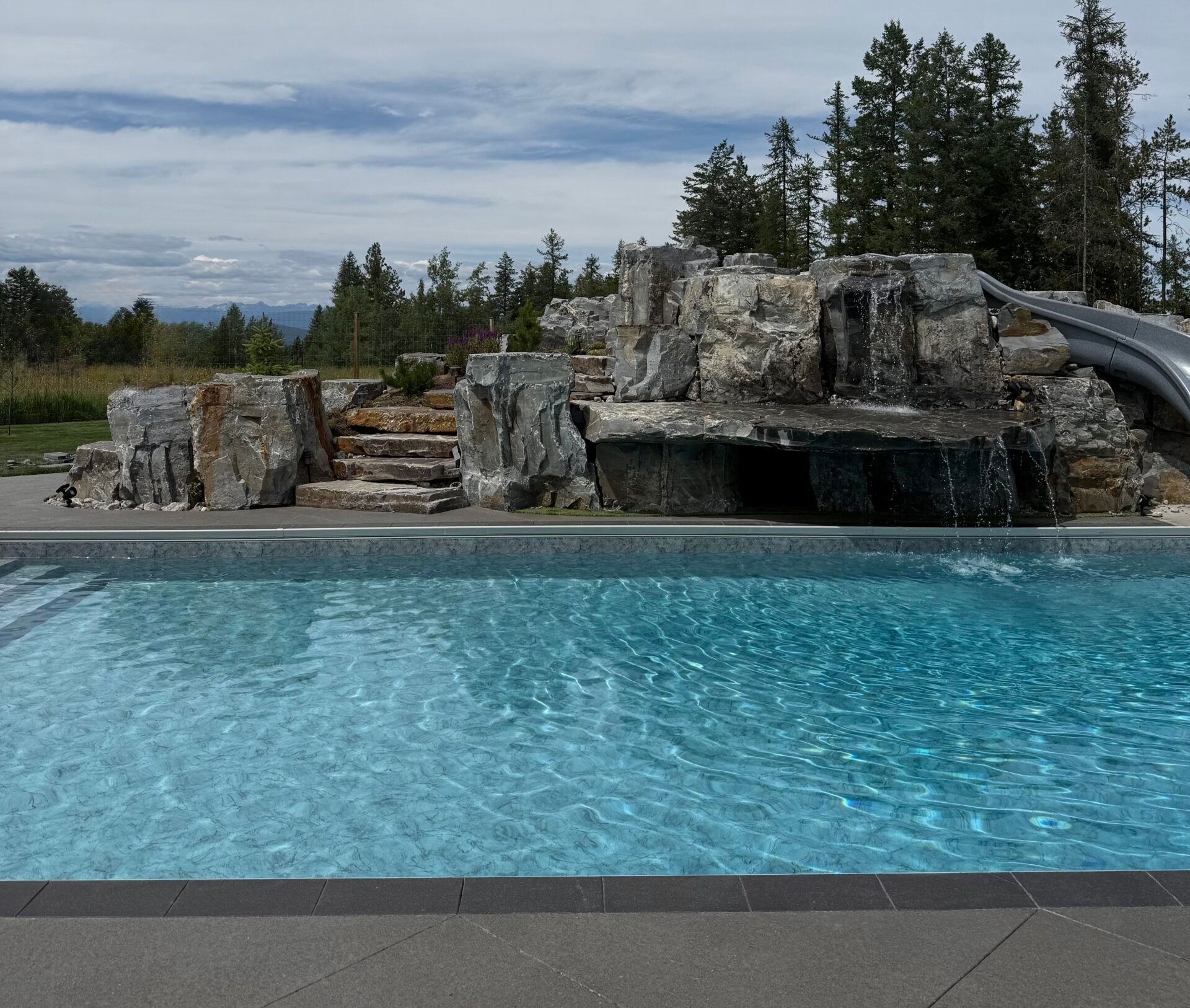 Blue swimming pool with rock waterfall and slide, backed by trees under a cloudy sky