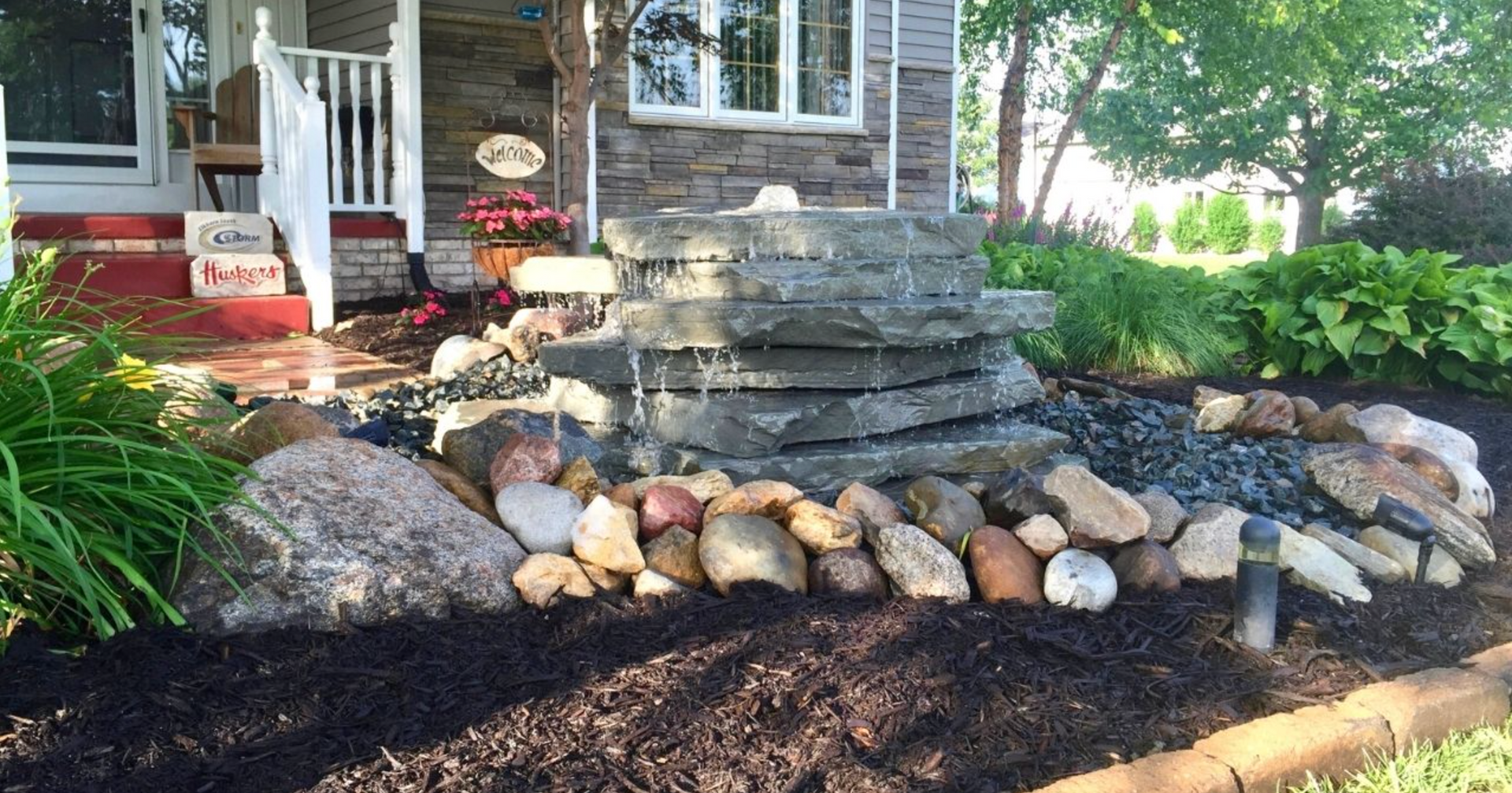 Stone steps lead to a front porch with potted flowers and a rock-lined garden bed in the yard