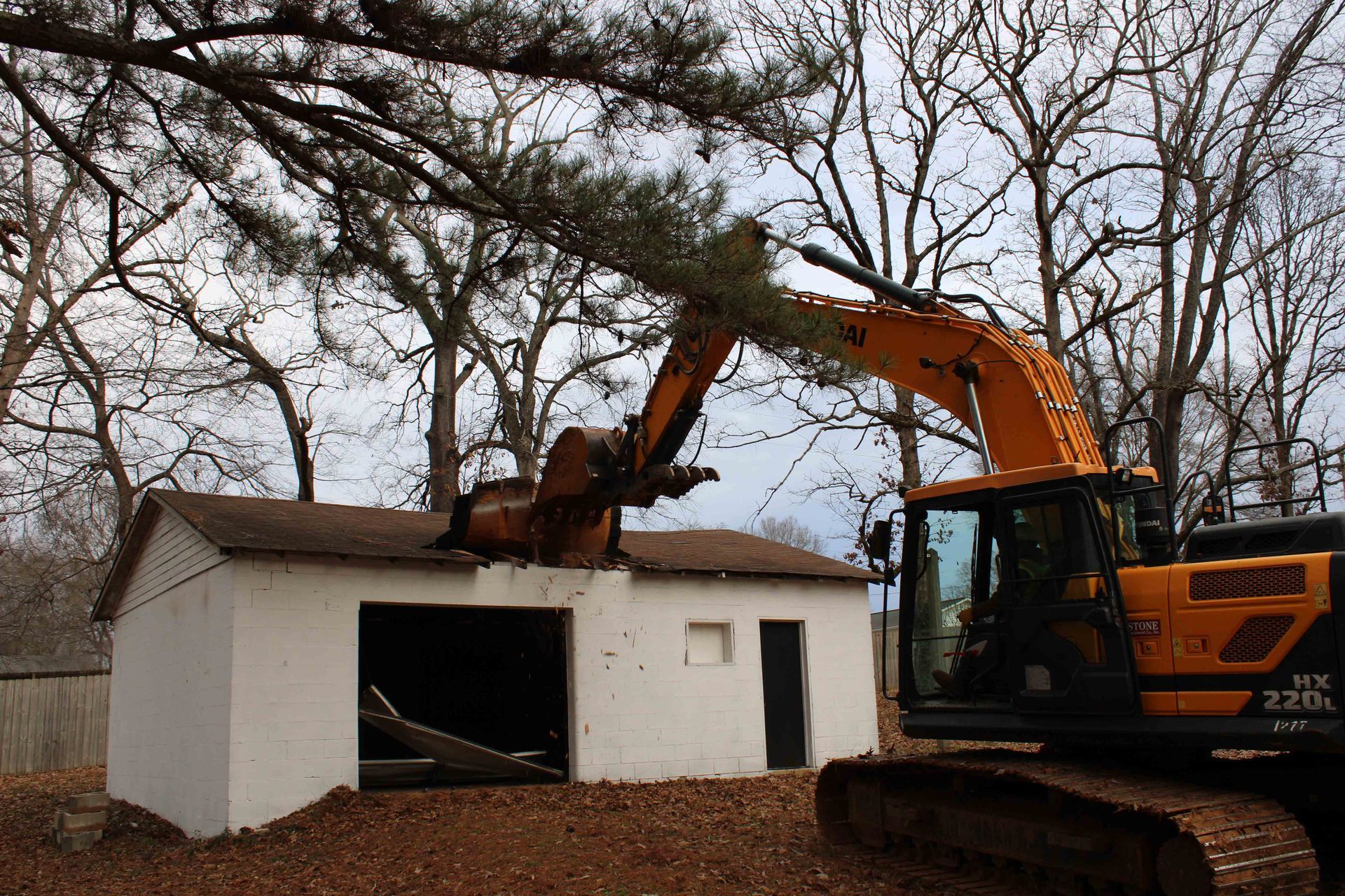 Excavator demolishing a small white house among bare trees in a wooded yard.