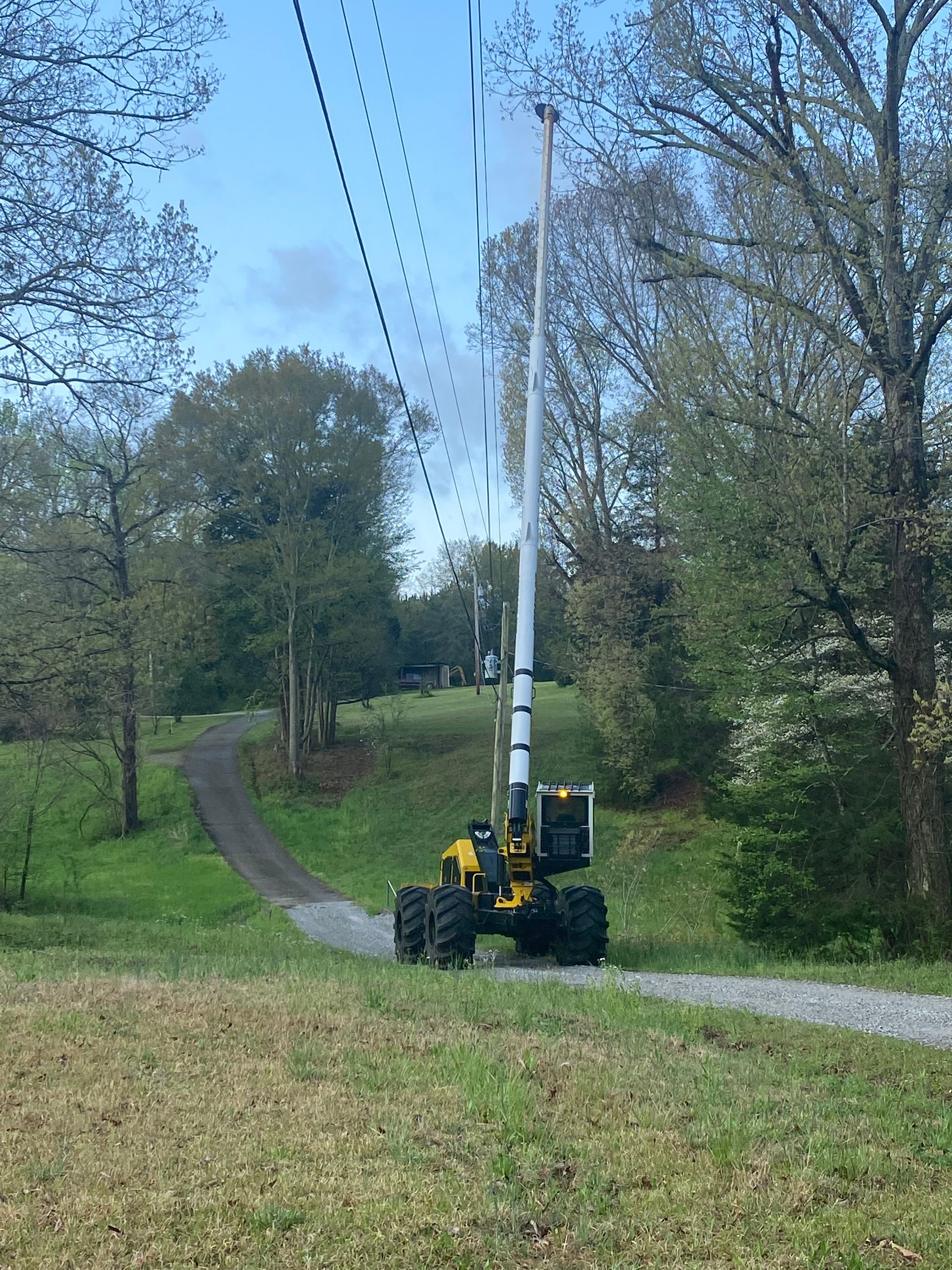 Crane lifting a tall white wind turbine blade on a rural dirt road amid trees and grass Crane lifting a tall white wind turbine blade on a rural dirt road amid trees and grass