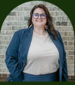 Person standing against a brick wall, wearing glasses, a blue blazer, and a light beige top. Person standing against a brick wall, wearing glasses, a blue blazer, and a light beige top.