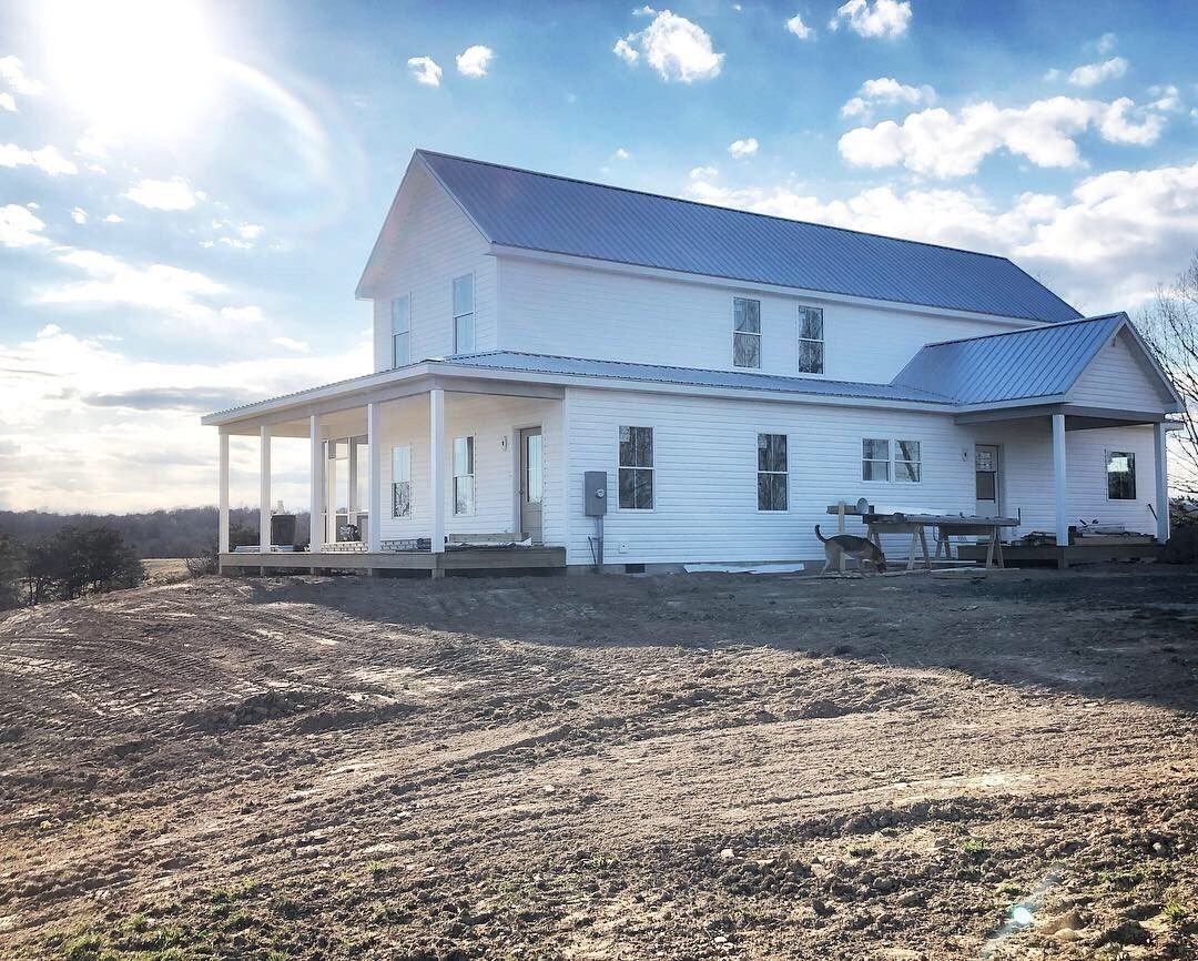 White farmhouse with wrap-around porch in a field under a sunny sky.