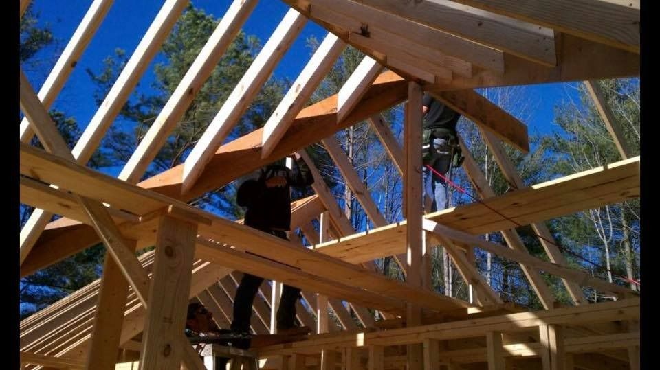 Construction workers building a wooden roof frame on a sunny day. Blue sky, wood beams, and tools are visible.