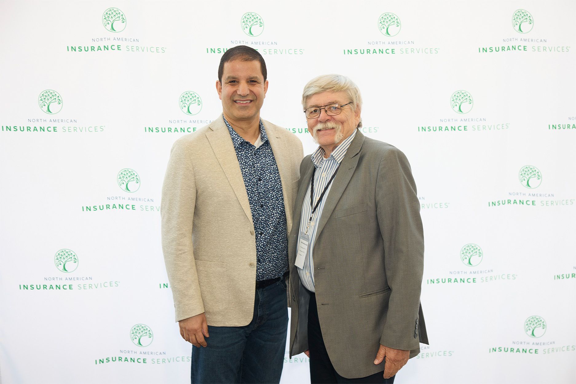 Two men in suits are standing next to each other in front of a white backdrop.