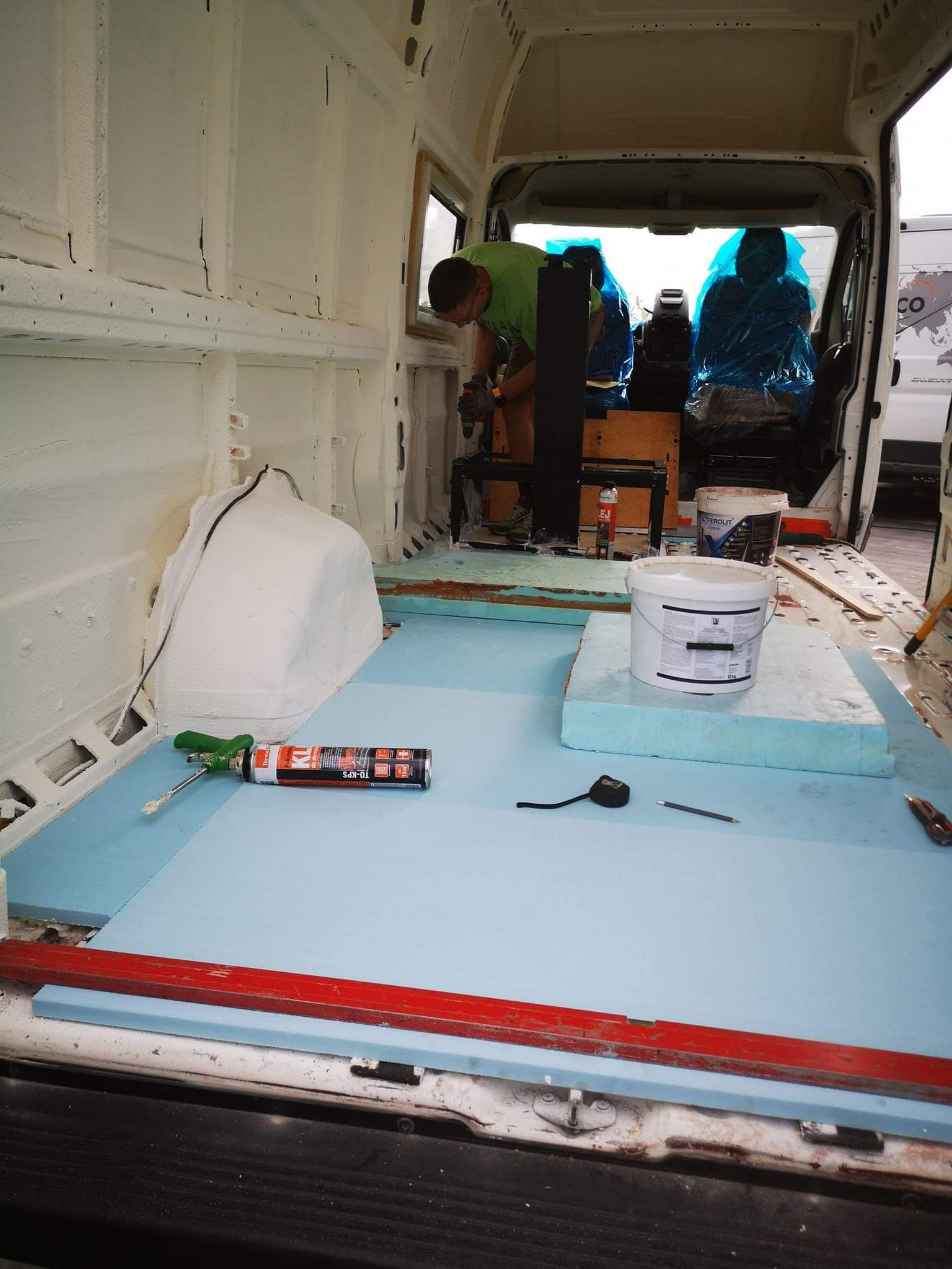 Inside a van being converted: man drilling into a blue foam floor, a bucket of white material, and tools.