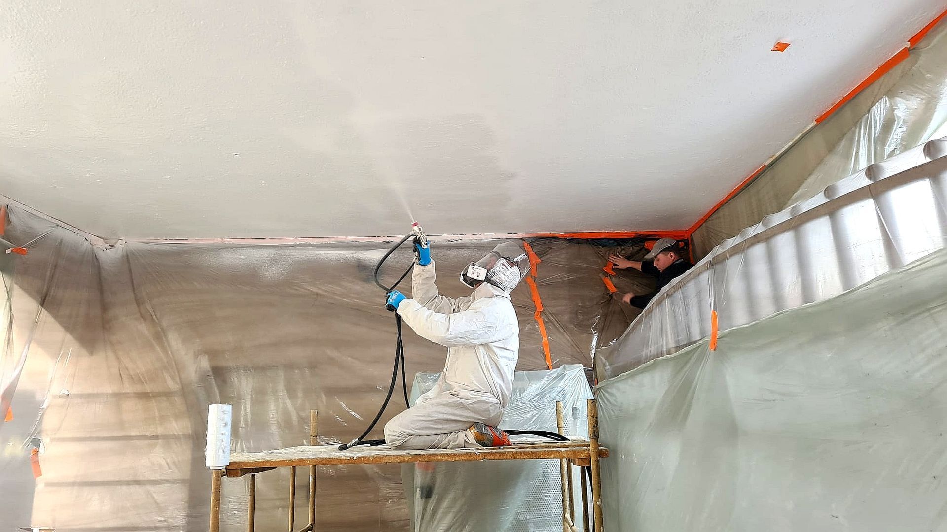 Person in protective suit sprays ceiling, room covered in plastic, orange tape on edges.