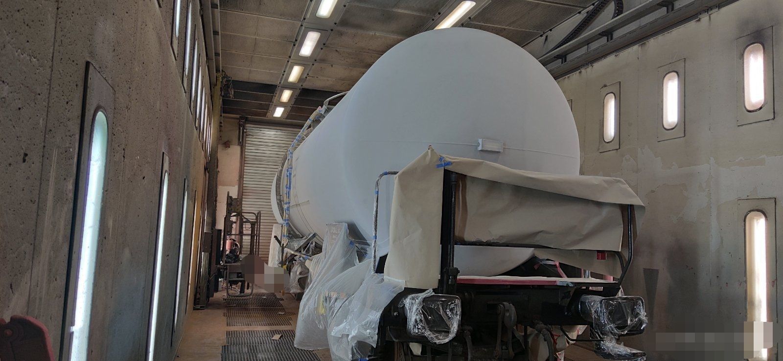 A large, white tank truck being painted inside a brightly lit industrial building.