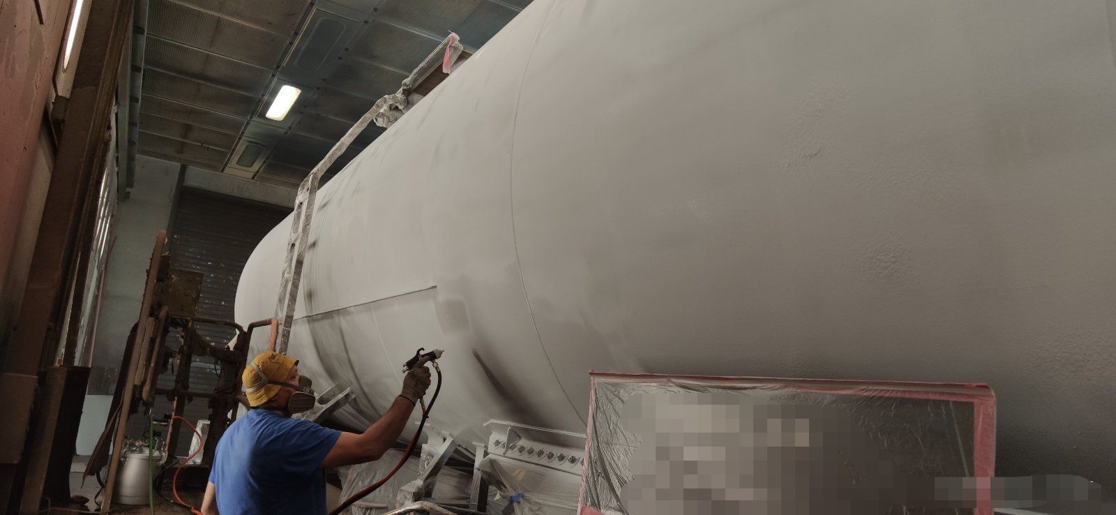 A worker sprays paint on a large cylindrical tank inside a warehouse.