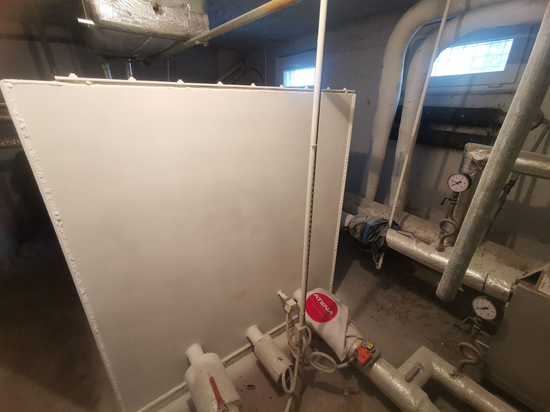 A mechanical room with white walls and pipes, featuring gauges, and a red and white object.