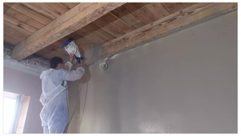 Person in protective suit sprays sealant on ceiling beams and wall indoors.