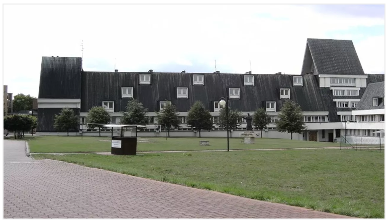Large gray building with a sloped roof, small windows, and a green lawn in front.