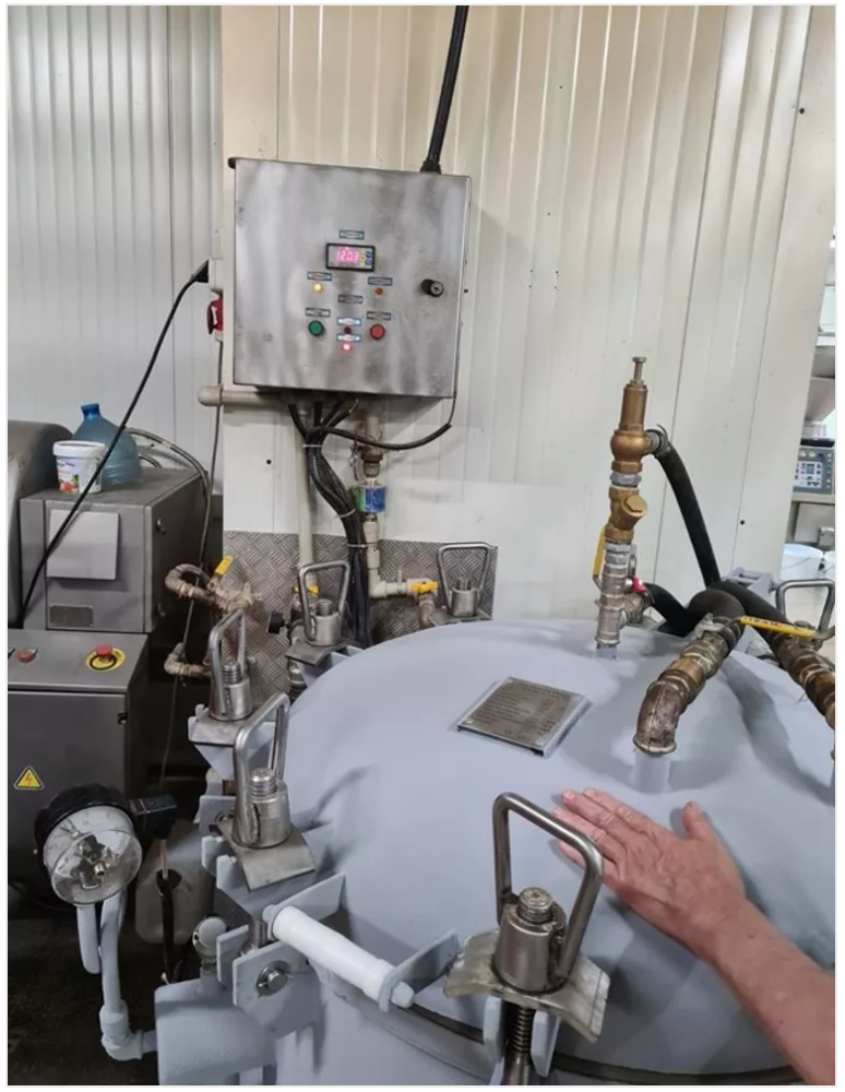 A person's hand touches a gray industrial machine with controls, valves, and piping in a white-walled room.