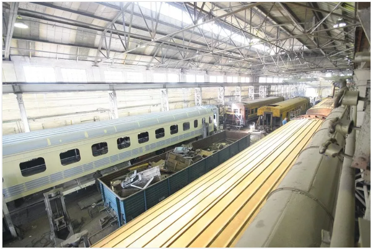 Inside a train car maintenance facility. Several train cars are undergoing repair, with workers and equipment visible.