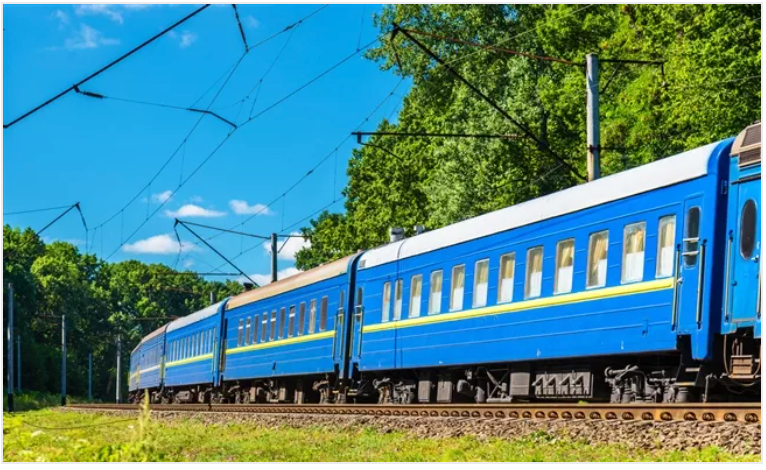 Blue passenger train on tracks, passing lush green trees under a clear blue sky.