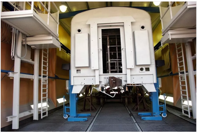 A train car in a maintenance bay, supported by blue jacks, surrounded by scaffolding.