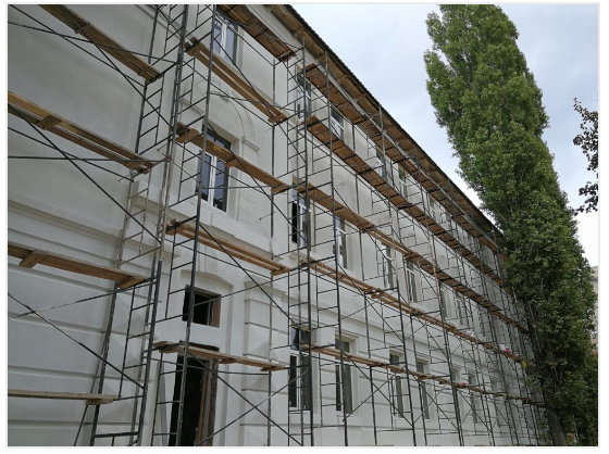 Building exterior with scaffolding, white walls, and a tall green tree.
