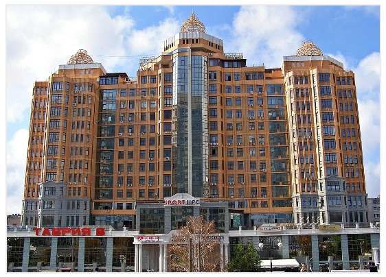 Tan-colored, multi-story building with shops at the base, topped with domed structures against a cloudy sky.