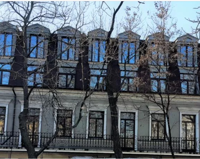 Gray building with many windows and a dark roof, trees in front.