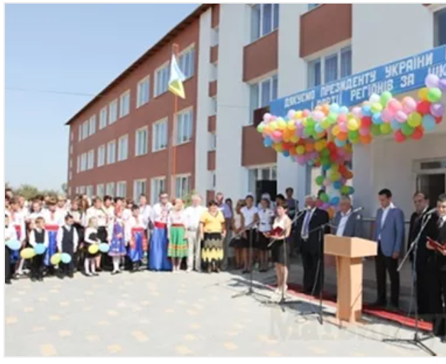School opening ceremony with people, balloons, flag, and dignitaries on a sunny day.