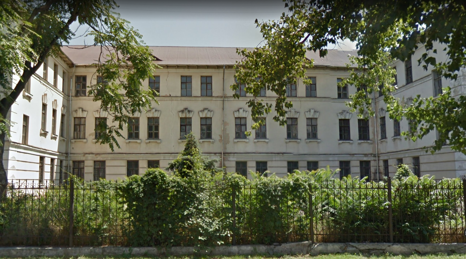 White multi-story building behind a wrought iron fence and lush green vegetation under a sunny sky.
