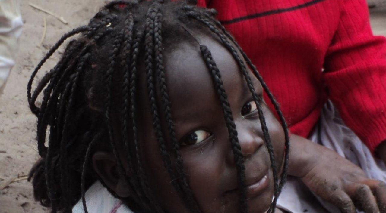 Child with braided hair leaning against a person in a red sweater, looking up at the camera.
