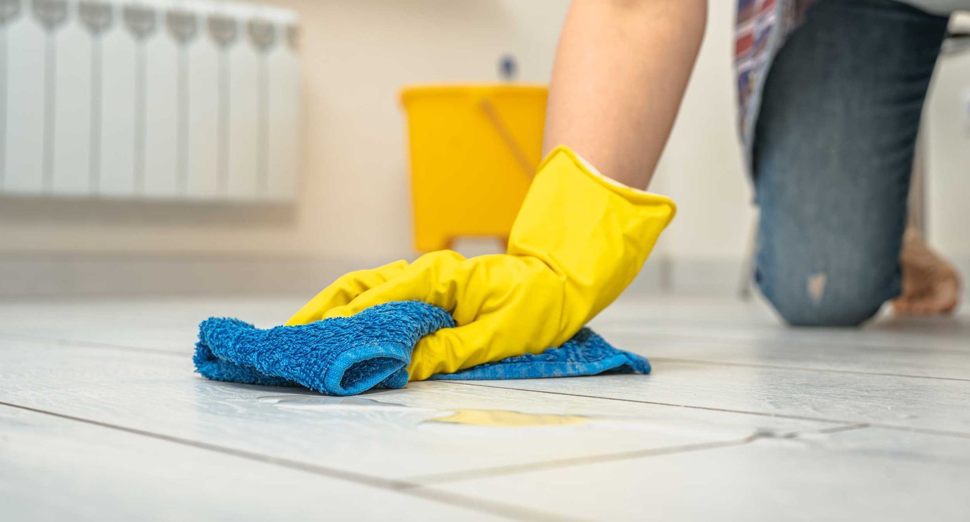 Person cleaning a white tile floor with a blue cloth and yellow gloves