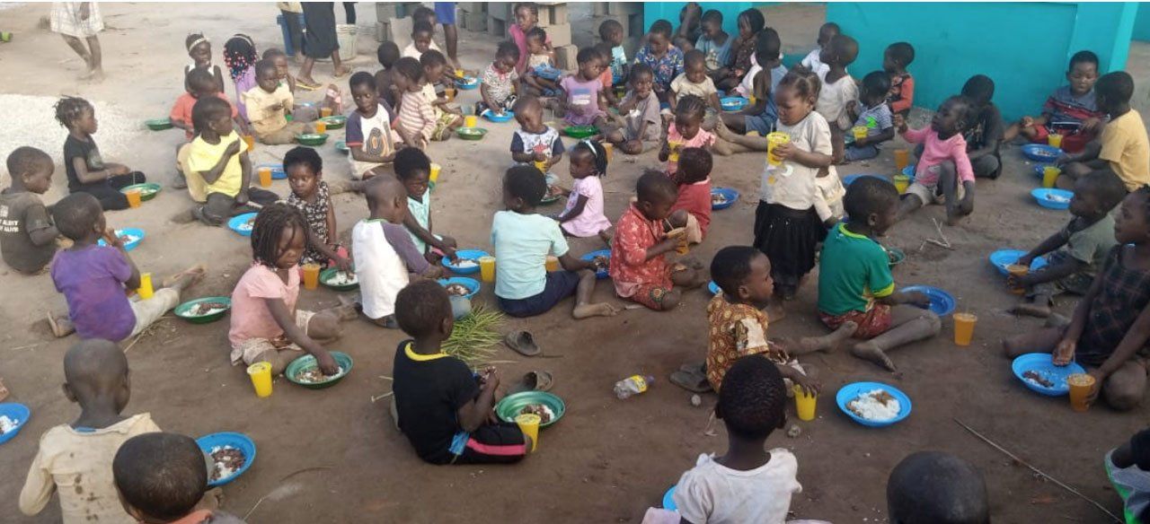 Children seated outdoors on the ground, eating from colorful plates and bowls in a group meal