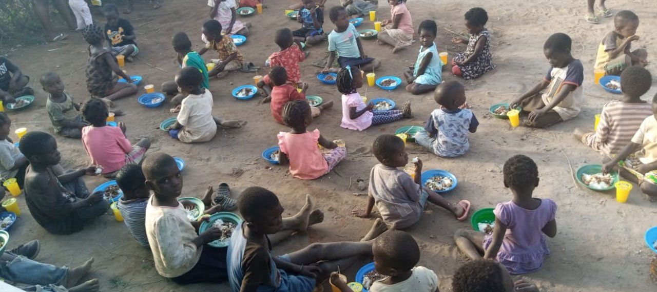 Children sitting outdoors in a circle, eating from bowls and plates on the ground.