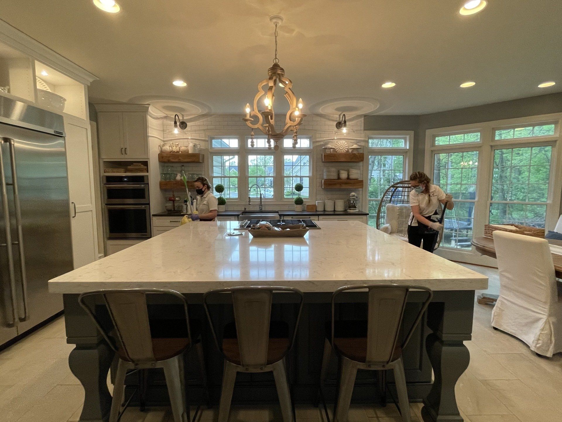 Bright kitchen with large island, chandelier, and a person standing by the windows.