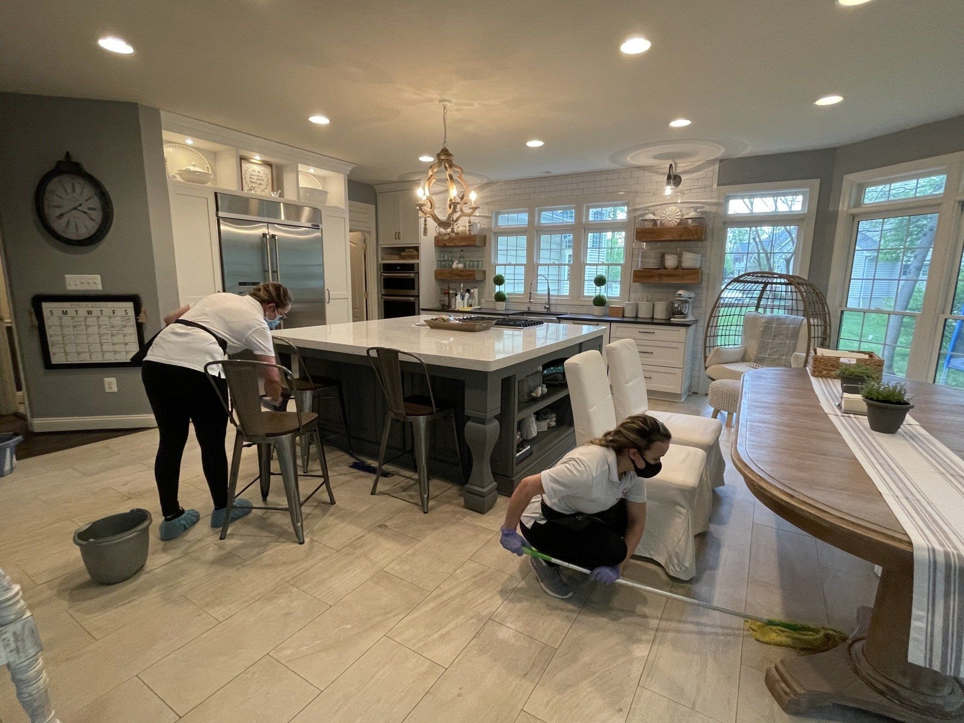 Two people cleaning a bright kitchen with white cabinets, island, and tiled floor.