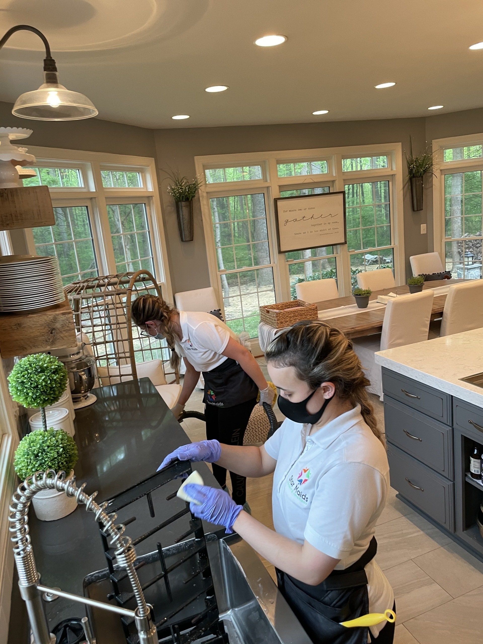 Two workers in masks clean a kitchen counter in a bright café with large windows.